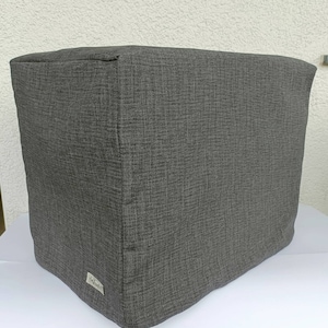 Puede incluir: Una funda de tela gris oscuro, posiblemente para un electrodoméstico o mueble. La funda tiene una superficie texturizada con un patrón tejido. Una pequeña etiqueta es visible en la esquina inferior. La funda es rectangular y parece estar bien ajustada.