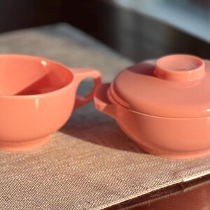 May include: A vintage peach-colored creamer and sugar bowl set. The creamer has a handle and a spout, while the sugar bowl has a lid and two small handles. Both pieces are made of a smooth, solid material.