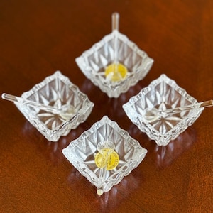 May include: Four clear glass leaf-shaped dishes with attached spoons. Each dish has a starburst pattern and a small spoon. One dish has a yellow sticker. The dishes are arranged on a dark brown surface.