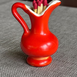 May include: A small, bright red ceramic jug with a curved handle and a white interior rim. The jug is filled with several wooden matches that have dark red tips, standing upright within the opening of the vessel.