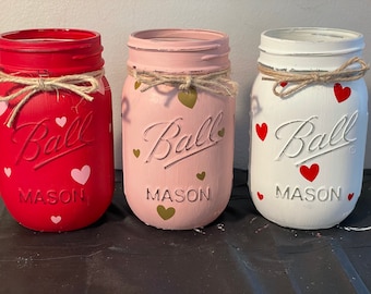 May include: Three mason jars painted in red, pink, and white with a heart pattern. Each jar has a brown twine bow and the text "Ball Mason" on the front.