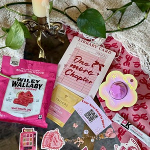 May include: A collection of pink and gold items, including a candle, snacks, tea, stickers, and a library card that says "One more Chapter." The items are arranged on a reflective surface, with a floral and bow pattern.