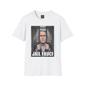 May include: A white short-sleeved t-shirt featuring a graphic print of a man in a suit behind prison bars. The man has gray hair and glasses, and his hands are gripping the bars. Below the image, the text reads JAIL FAUCI in bold white letters.