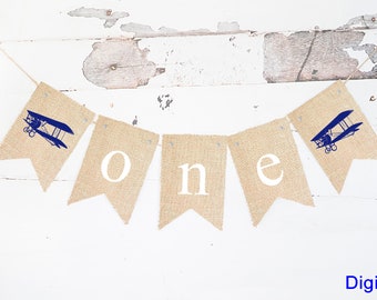 Könnte beinhalten: Ein Banner aus Jute mit dem Wort "one" in weißen Buchstaben und zwei blauen Flugzeugen an den Enden. Das Banner ist mit einer Schnur befestigt und perfekt für eine erste Geburtstagsfeier.