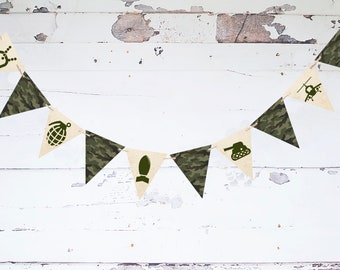 May include: A green and tan camouflage patterned bunting with military themed symbols including a tank, grenade, helicopter, and dog tag.