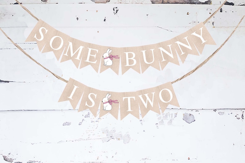 以下が含まれることがあります： 白い文字で「Some Bunny Is Two」と書かれた麻の横断幕。横断幕は、ピンクのリボンをつけた白いウサギ2匹で飾られています。