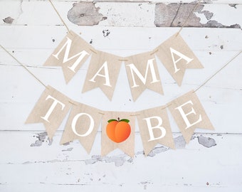 May include: A burlap banner with the words "Mama To Be" in white letters. A peach with a green stem is between the words "To" and "Be".