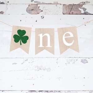 Puede incluir: Una pancarta de arpillera con un trébol de cuatro hojas de purpurina verde y la palabra "one" en letras blancas. La pancarta está colgada sobre un fondo de madera blanca.