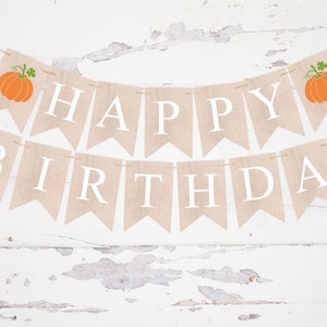 Puede incluir: Una pancarta de arpillera con las palabras "Happy Birthday" en letras blancas. La pancarta está decorada con dos calabazas naranjas con tallos y hojas verdes.