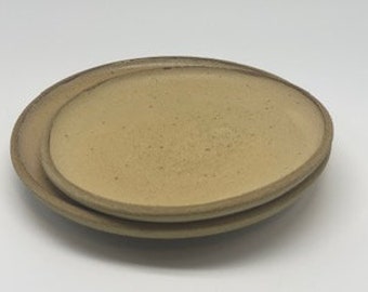 May include: Two beige ceramic plates with a slightly irregular, rounded shape. The plates are stacked on top of each other.