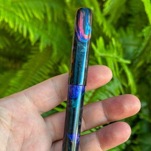 May include: A colorful pen with a swirling pattern of blue, purple, orange, and black. The pen is held in a hand against a backdrop of green foliage. The pen's design is visible on the cap and body.
