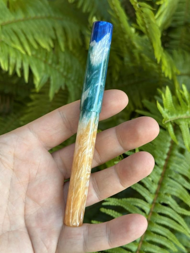 May include: A handcrafted pen with a unique design. The pen features a gradient of colours, starting with a blue top, transitioning to teal and white, and ending with a golden-yellow base. The pen is held against a backdrop of green foliage.