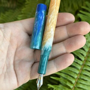 May include: A handcrafted fountain pen with a vibrant design. The pen features a gold nib and a barrel that transitions from yellow to white and teal. The cap is blue and teal. The pen is held against a backdrop of green foliage.