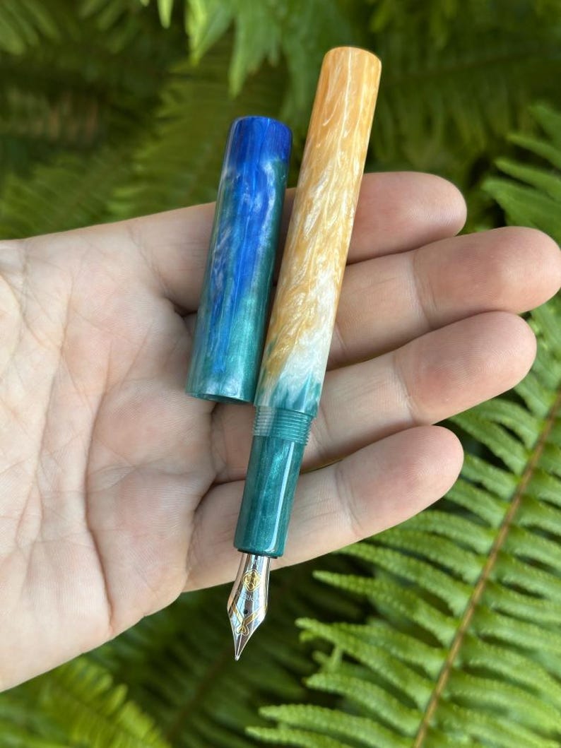 May include: A handcrafted fountain pen with a vibrant design. The pen features a gold nib and a barrel that transitions from yellow to white and teal. The cap is blue and teal. The pen is held against a backdrop of green foliage.