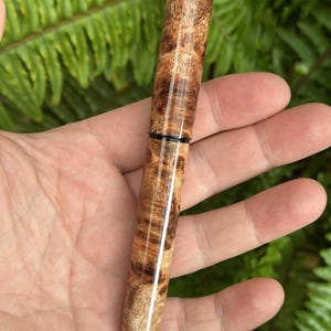 Puede incluir: Un bol&iacute;grafo de madera hecho a mano con un patr&oacute;n de veta de madera de ra&iacute;z natural. El bol&iacute;grafo es cil&iacute;ndrico con un acabado pulido, con una banda oscura cerca del centro. El color de la madera var&iacute;a de marr&oacute;n claro a oscuro, con patrones intrincados.