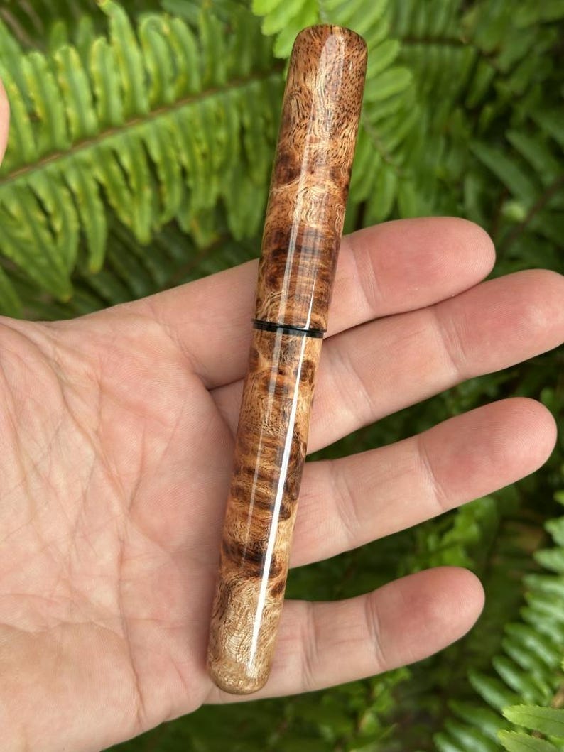 Puede incluir: Un bol&iacute;grafo de madera hecho a mano con un patr&oacute;n de veta de madera de ra&iacute;z natural. El bol&iacute;grafo es cil&iacute;ndrico con un acabado pulido, con una banda oscura cerca del centro. El color de la madera var&iacute;a de marr&oacute;n claro a oscuro, con patrones intrincados.