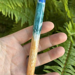 May include: A handcrafted pen with a unique design. The pen features a color gradient, transitioning from blue at the top, through shades of green and white, to a golden-yellow at the base. The pen is held against a backdrop of green foliage.