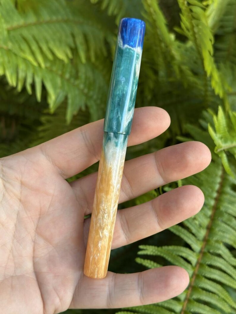 May include: A handcrafted pen with a unique design. The pen features a color gradient, transitioning from blue at the top, through shades of green and white, to a golden-yellow at the base. The pen is held against a backdrop of green foliage.