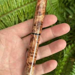 Puede incluir: Un bol&iacute;grafo de madera con un patr&oacute;n natural de vetas de madera de ra&iacute;z. El bol&iacute;grafo es cil&iacute;ndrico con una punta redondeada y una banda negra cerca del centro. El bol&iacute;grafo se sostiene contra un fondo de follaje verde.