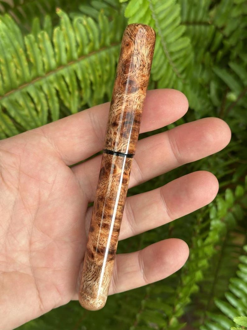 Puede incluir: Un bol&iacute;grafo de madera con un patr&oacute;n natural de vetas de madera de ra&iacute;z. El bol&iacute;grafo es cil&iacute;ndrico con una punta redondeada y una banda negra cerca del centro. El bol&iacute;grafo se sostiene contra un fondo de follaje verde.