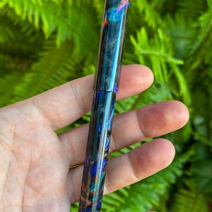 May include: A colorful pen with a swirling pattern of blues, greens, and oranges. The pen has a glossy finish and a cylindrical shape. The pen is held in a hand against a backdrop of green foliage.