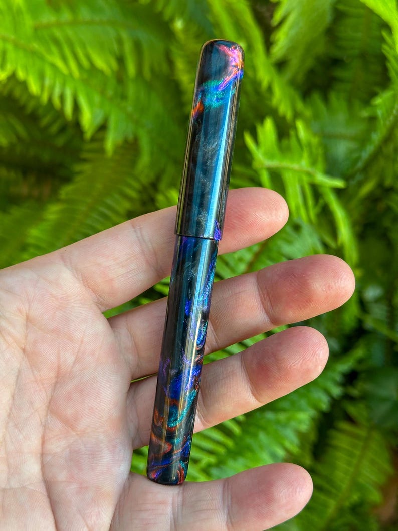May include: A colorful pen with a swirling pattern of blues, greens, and oranges. The pen has a glossy finish and a cylindrical shape. The pen is held in a hand against a backdrop of green foliage.