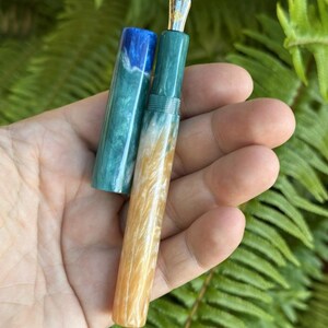 May include: A handcrafted fountain pen with a gold nib and a colorful resin body. The pen features a teal, blue, and gold marbled design. The cap is off, revealing the nib and the pen's internal components. The pen is held in a hand against a backdrop of green foliage.