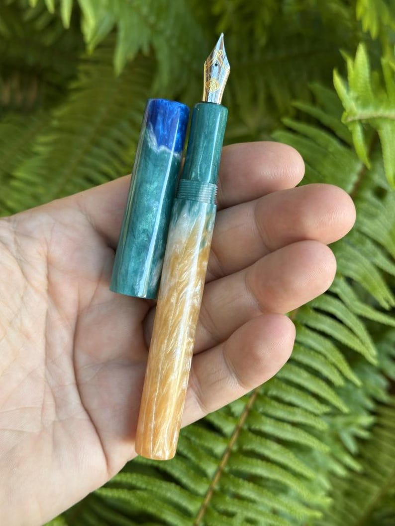 May include: A handcrafted fountain pen with a gold nib and a colorful resin body. The pen features a teal, blue, and gold marbled design. The cap is off, revealing the nib and the pen's internal components. The pen is held in a hand against a backdrop of green foliage.