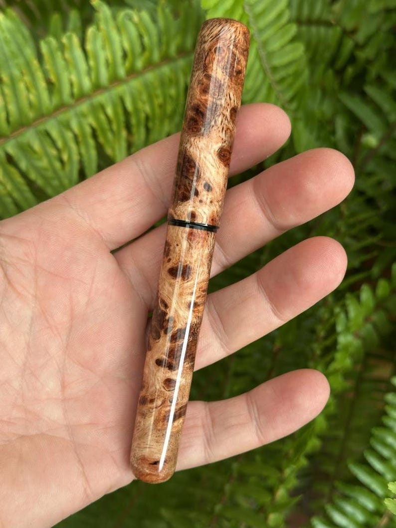 Puede incluir: Un bol&iacute;grafo de madera artesanal con un patr&oacute;n de veta de madera de burl &uacute;nico. El bol&iacute;grafo presenta un acabado pulido, mostrando los colores naturales de la madera, con tonos marr&oacute;n oscuro y marr&oacute;n claro. Una banda negra acent&uacute;a el dise&ntilde;o del bol&iacute;grafo.