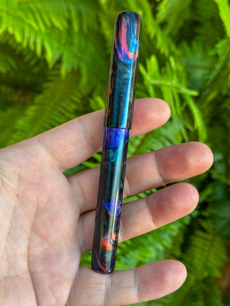 May include: A colorful pen with a swirling pattern of blue, purple, orange, and black. The pen is held in a hand against a backdrop of green foliage. The pen's design is visible on the cap and body.