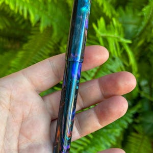 May include: A colorful fountain pen with a swirling pattern of blues, greens, oranges, and purples. The pen has a glossy finish and a cylindrical shape. The pen is held in a hand against a backdrop of green foliage.