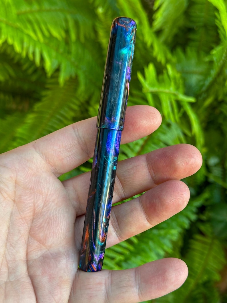 May include: A colorful fountain pen with a swirling pattern of blues, greens, oranges, and purples. The pen has a glossy finish and a cylindrical shape. The pen is held in a hand against a backdrop of green foliage.