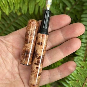 Puede incluir: Una pluma estilogr&aacute;fica artesanal con cuerpo y capuch&oacute;n de madera, con un patr&oacute;n de vetas de madera de burl &uacute;nico en tonos marrones y beige. La pluma tiene una secci&oacute;n de agarre negra y un plum&iacute;n plateado. Se ha quitado el capuch&oacute;n.