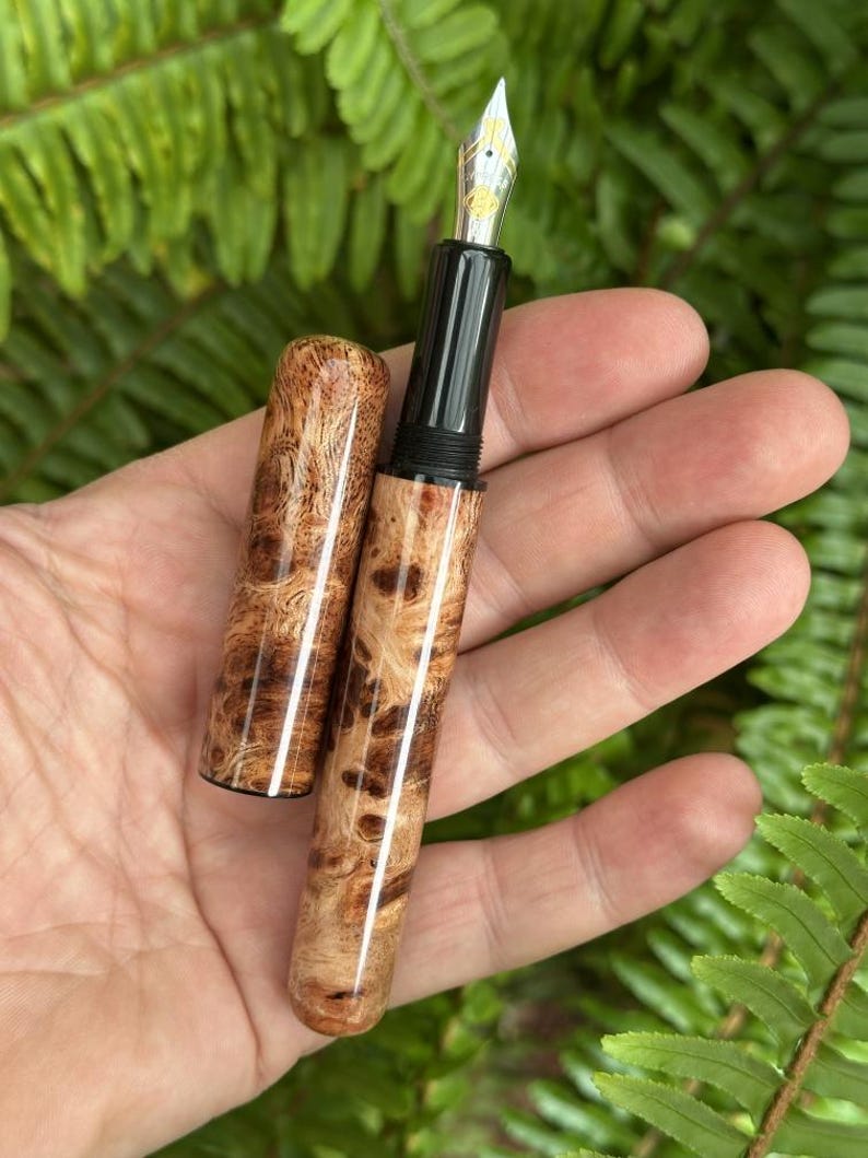 Puede incluir: Una pluma estilogr&aacute;fica artesanal con cuerpo y capuch&oacute;n de madera, con un patr&oacute;n de vetas de madera de burl &uacute;nico en tonos marrones y beige. La pluma tiene una secci&oacute;n de agarre negra y un plum&iacute;n plateado. Se ha quitado el capuch&oacute;n.