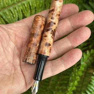 Puede incluir: Una pluma estilogr&aacute;fica artesanal con cuerpo y capuch&oacute;n de madera, con un patr&oacute;n de vetas de madera de burl &uacute;nico en tonos marrones y beige. La pluma tiene una secci&oacute;n de agarre negra y un plum&iacute;n plateado con detalles dorados. Se ha quitado el capuch&oacute;n.