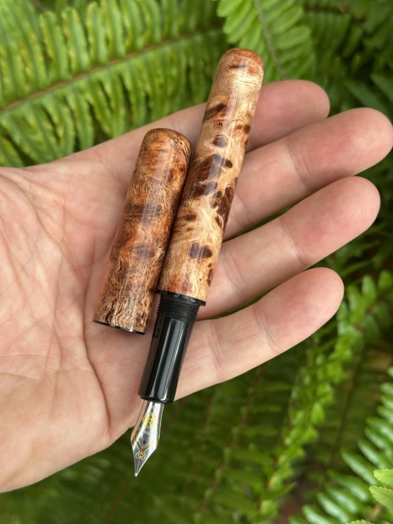 Puede incluir: Una pluma estilogr&aacute;fica artesanal con cuerpo y capuch&oacute;n de madera, con un patr&oacute;n de vetas de madera de burl &uacute;nico en tonos marrones y beige. La pluma tiene una secci&oacute;n de agarre negra y un plum&iacute;n plateado con detalles dorados. Se ha quitado el capuch&oacute;n.