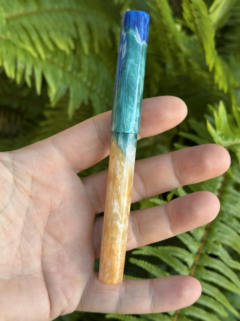 May include: A handcrafted pen with a unique design. The pen features a blue, teal, and white upper barrel, transitioning to a tan and white lower barrel. The pen is held against a backdrop of green foliage.