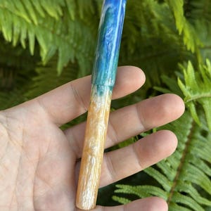 May include: A handcrafted pen with a unique design. The pen's barrel transitions from a deep blue at the top, through shades of teal and green, to a warm, sandy beige at the base. The pen is held against a backdrop of green foliage.