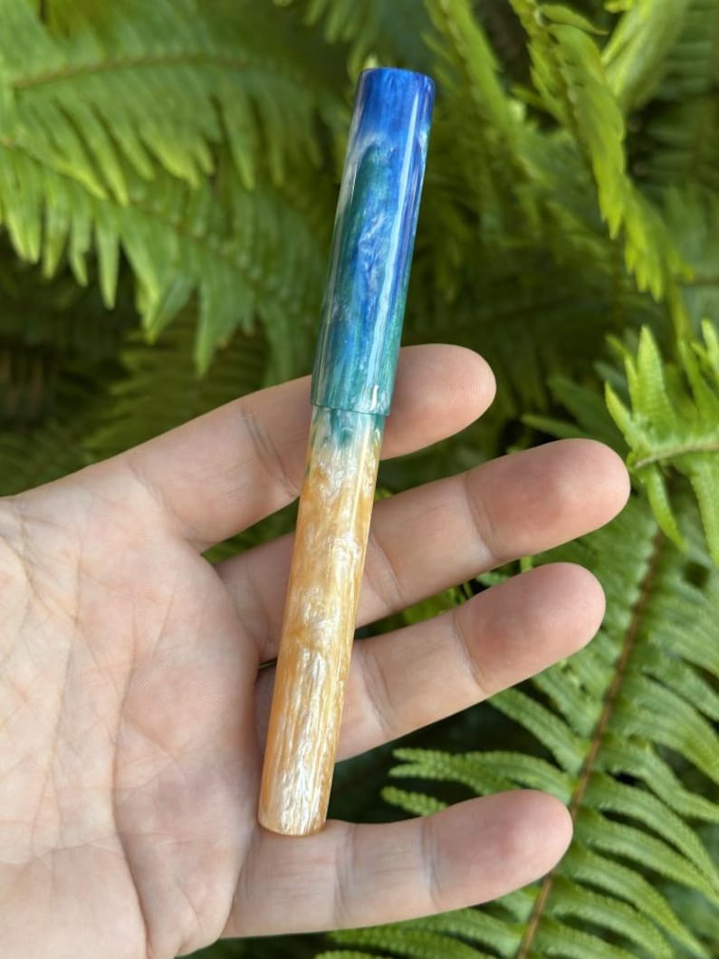May include: A handcrafted pen with a unique design. The pen's barrel transitions from a deep blue at the top, through shades of teal and green, to a warm, sandy beige at the base. The pen is held against a backdrop of green foliage.