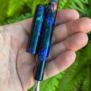 May include: A handcrafted fountain pen with a vibrant, swirling pattern of blue, teal, and orange. The pen features a gold-colored nib and a matching cap. The pen is held against a backdrop of green foliage.