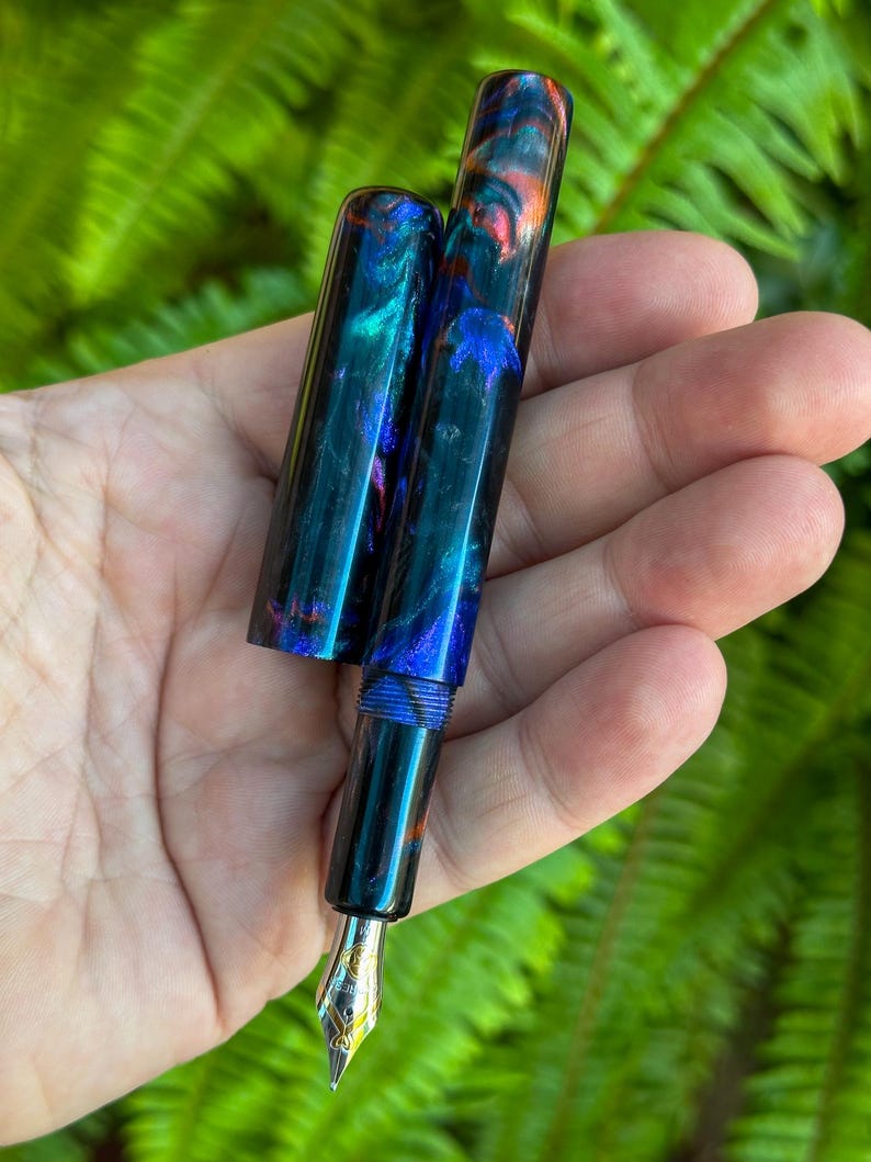 May include: A handcrafted fountain pen with a vibrant, swirling pattern of blue, teal, and orange. The pen features a gold-colored nib and a matching cap. The pen is held against a backdrop of green foliage.