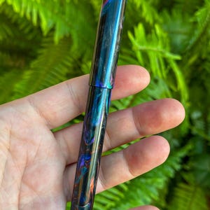 May include: A handcrafted fountain pen with a swirling pattern of blue, teal, orange, and purple. The pen has a glossy finish and a sleek, cylindrical design. The pen is held in a hand against a backdrop of green foliage.