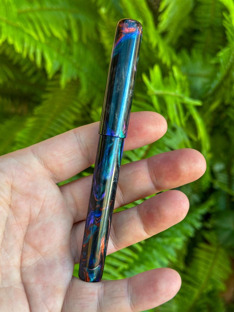 May include: A handcrafted fountain pen with a swirling pattern of blue, teal, orange, and purple. The pen has a glossy finish and a sleek, cylindrical design. The pen is held in a hand against a backdrop of green foliage.