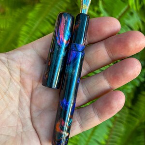 May include: A handcrafted fountain pen with a vibrant, swirling pattern of blue, purple, and orange hues. The pen features a gold-colored nib with intricate detailing. The cap is off, revealing the pen's design. The pen is held in a hand against a backdrop of green foliage.