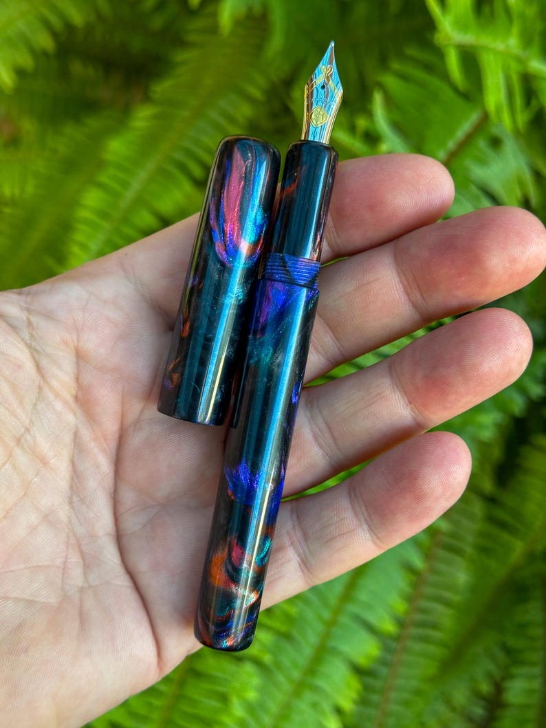 May include: A handcrafted fountain pen with a vibrant, swirling pattern of blue, purple, and orange hues. The pen features a gold-colored nib with intricate detailing. The cap is off, revealing the pen's design. The pen is held in a hand against a backdrop of green foliage.