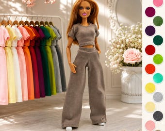 Può includere: Una bambola alla moda con lunghi capelli color ramato indossa un completo coordinato color tortora composto da un top corto a maniche corte e pantaloni a gamba larga. La bambola è posizionata davanti a un appendiabiti con vari outfit colorati.