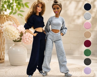 Può includere: Due bambole indossano completi lounge a due pezzi abbinati composti da top corti a maniche lunghe e pantaloni della tuta a vita alta con polsini elastici. Una bambola indossa un completo blu scuro, l'altra un completo grigio melange.