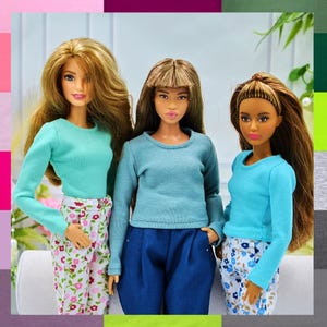 May include: Three dolls wearing long-sleeved shirts in different shades of blue. The dolls are wearing pants with floral patterns. The dolls are posed against a white background.