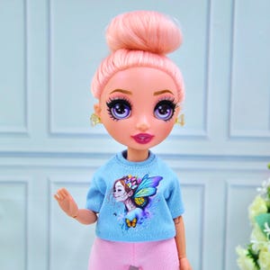Puede incluir: Una muñeca con cabello rosado recogido en un moño, que lleva una camiseta azul claro con un diseño de hada y pantalones rosas. La muñeca tiene ojos grandes de color púrpura, labios rosados y pequeños pendientes amarillos. La piel de la muñeca es de color melocotón claro.
