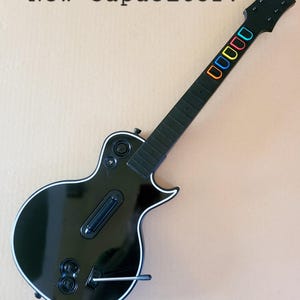 May include: Black guitar-shaped game controller with white trim. The fretboard has colorful buttons. The text "New Capacitor!" is above the guitar. The headstock has the "Gibson" logo.